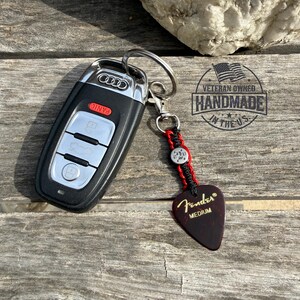 Può includere: Chiave per auto Audi nera con portachiavi a plettro per chitarra. Il portachiavi ha un cordino rosso e nero con un bottone in metallo e un plettro Fender rosso scuro. Il plettro dice "Fender MEDIUM". L'immagine include anche il testo "Veteran Owned Handmade in the US."