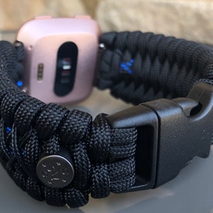 Puede incluir: Pulsera de reloj de paracord negro con hebilla negra y bot&oacute;n de metal plateado. La pulsera est&aacute; tejida con detalles azules.