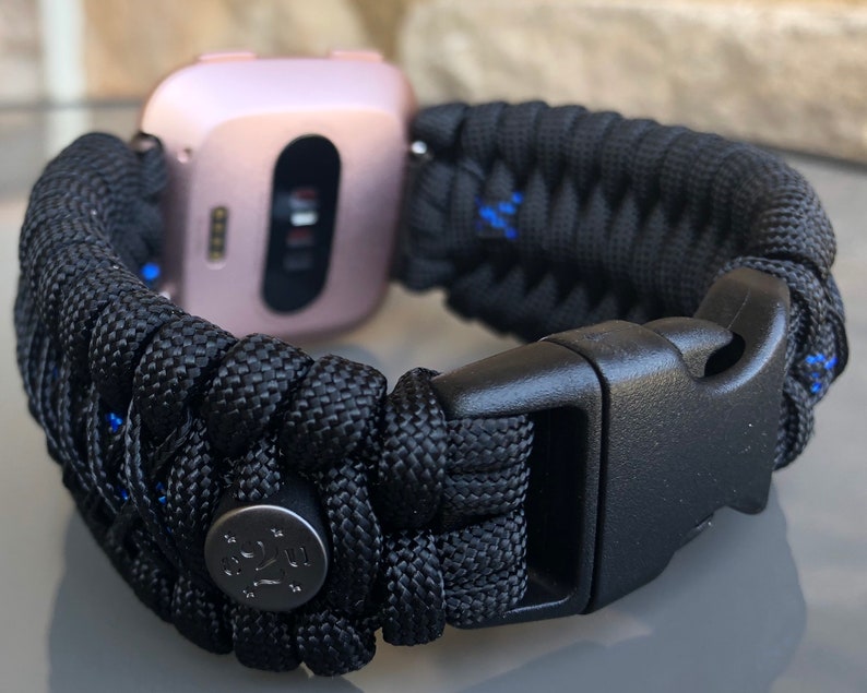 Puede incluir: Pulsera de reloj de paracord negro con hebilla negra y bot&oacute;n de metal plateado. La pulsera est&aacute; tejida con detalles azules.