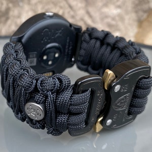 May include: A black smartwatch featuring a woven paracord band with a textured, knotted design. The band includes a metallic silver bead with star engravings and a black metal Cobra buckle with gold-toned release levers for a secure closure.