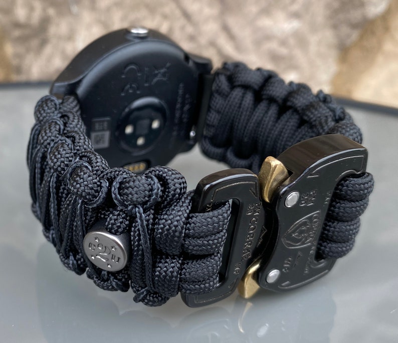 May include: A black smartwatch featuring a woven paracord band with a textured, knotted design. The band includes a metallic silver bead with star engravings and a black metal Cobra buckle with gold-toned release levers for a secure closure.