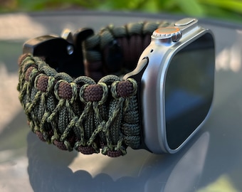 Op de afbeelding: Een zilveren Apple Watch met een groen en bruin paracord-bandje. Het bandje is geweven met een patroon van groene en bruine koorden.
