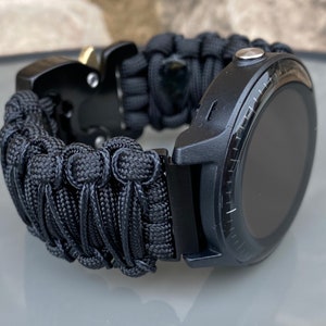 May include: A black smartwatch featuring a round display and a thick, woven black paracord strap. The band is secured with a black metal side-release buckle that includes a small gold-toned accent piece. The watch case is matte black with a silver button.