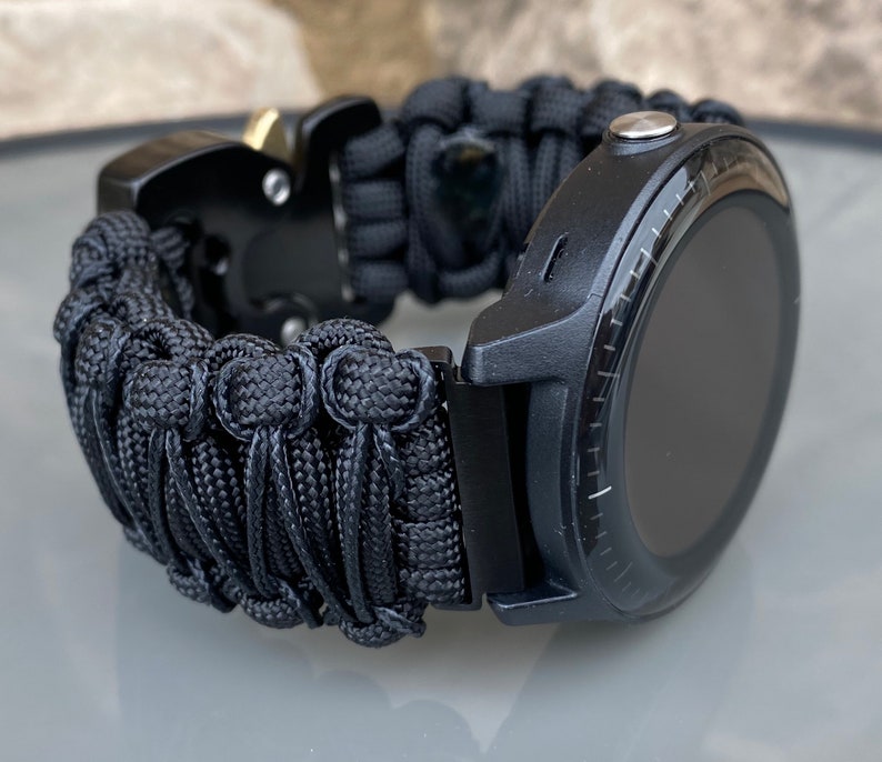 May include: A black smartwatch featuring a round display and a thick, woven black paracord strap. The band is secured with a black metal side-release buckle that includes a small gold-toned accent piece. The watch case is matte black with a silver button.