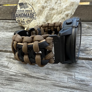 Op de afbeelding: Een zwart horloge met een op maat gemaakte paracord band in bruin en zwart. De band is ingewikkeld geweven. De afbeelding bevat een logo "Veteran Owned Handmade in the U.S.".