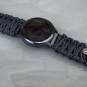 May include: A black smartwatch featuring a round face and a woven black paracord strap. The strap is secured with a black metal buckle and includes a small silver-toned circular accent piece. The watch face is dark and circular with a thin bezel.