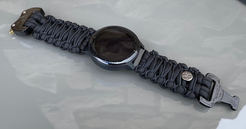 May include: A black smartwatch featuring a round face and a woven black paracord strap. The strap is secured with a black metal buckle and includes a small silver-toned circular accent piece. The watch face is dark and circular with a thin bezel.