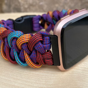 Puede incluir: Un reloj inteligente Fitbit de color rosa dorado con una correa trenzada de paracord de colores. La correa est&aacute; hecha de paracord rojo, azul, amarillo y turquesa.