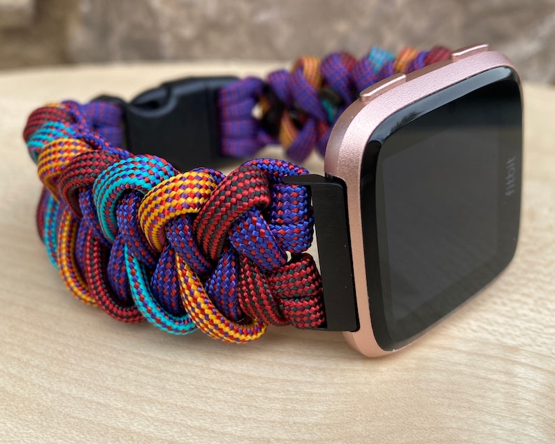 Puede incluir: Un reloj inteligente Fitbit de color rosa dorado con una correa trenzada de paracord de colores. La correa est&aacute; hecha de paracord rojo, azul, amarillo y turquesa.