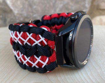 May include: A black smartwatch with a red, white, and black paracord strap. The strap is woven in a complex pattern.