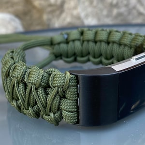 Op de afbeelding: Een groene paracord armband met een zwarte en zilveren fitness tracker bevestigd. De armband is gemaakt met een geweven paracord patroon.