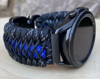 May include: Black smartwatch with a black and gray paracord band featuring a blue stripe. The watch has a black bezel and a digital display. The band is made of woven nylon cord.