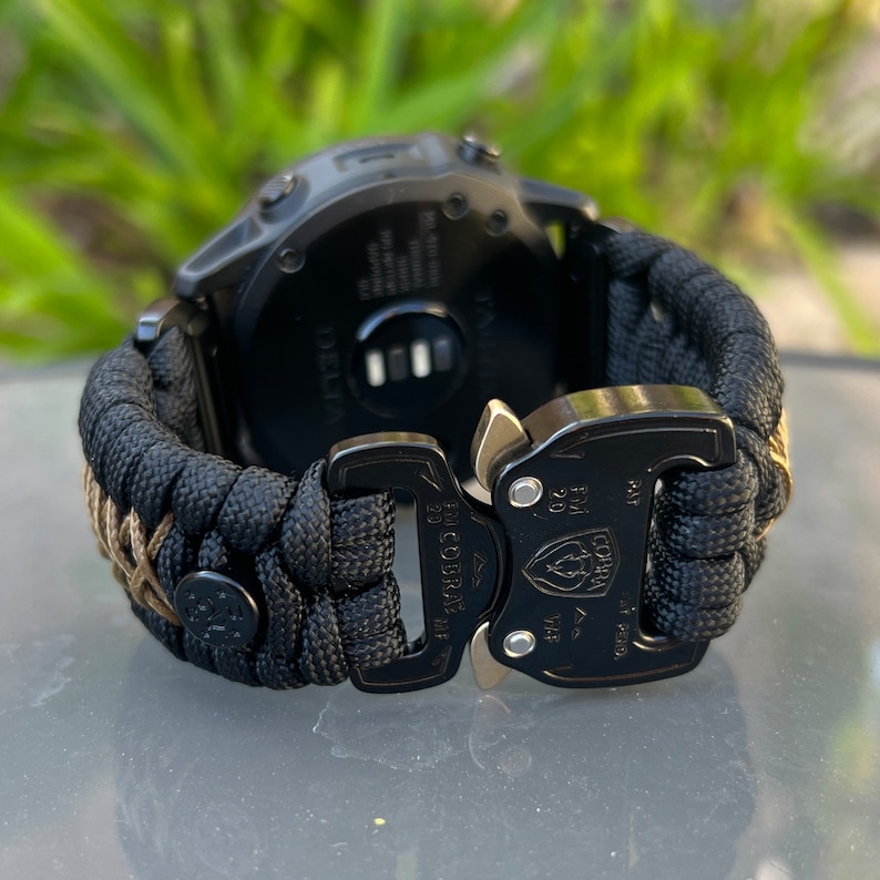 May include: Black watch band with a black buckle and a black and tan paracord strap. The buckle has the text "20 MM COBRA-DF" and "FOR 20 MM" on it.