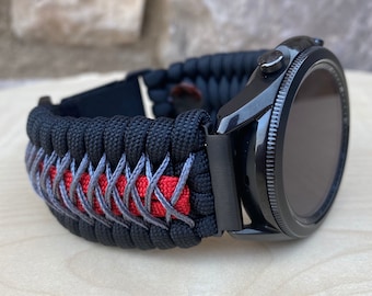 May include: Black smartwatch with a black and gray paracord band featuring a red accent. The watch has a black bezel and a dark screen. The band is intricately woven, showcasing a durable and stylish design.