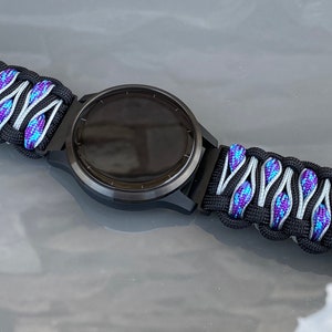 May include: A black smartwatch with a paracord watch band featuring a woven pattern of black, silver, and iridescent blue-purple cord. The band includes a black plastic side-release buckle and a small circular silver-toned metal accent piece.