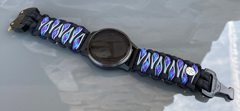 May include: A black smartwatch with a paracord watch band featuring a woven pattern of black, silver, and iridescent blue-purple cord. The band includes a black plastic side-release buckle and a small circular silver-toned metal accent piece.