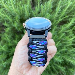May include: A smartwatch featuring a black circular face and a custom paracord watch band. The band is constructed from black cord woven with a central pattern of grey cord and a vibrant blue and purple speckled cord, creating a textured design.