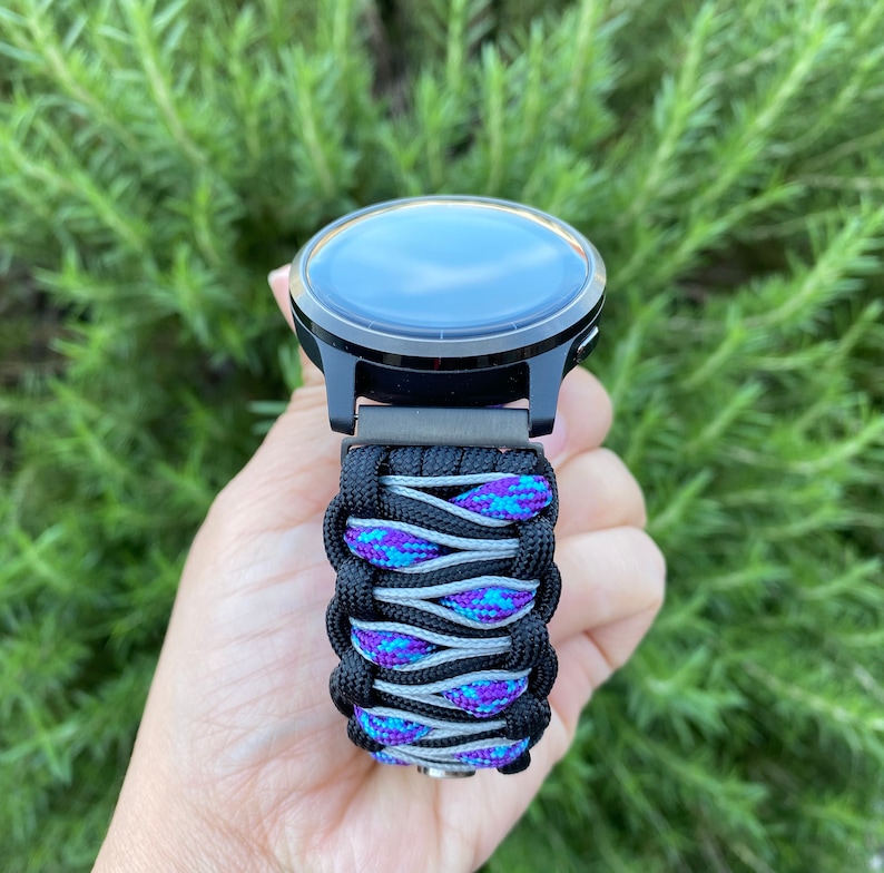 May include: A smartwatch featuring a black circular face and a custom paracord watch band. The band is constructed from black cord woven with a central pattern of grey cord and a vibrant blue and purple speckled cord, creating a textured design.