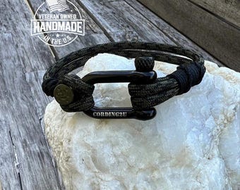 Puede incluir: Pulsera de paracord negro con un cierre de metal negro. La pulsera tiene un patrón de camuflaje verde y el cierre tiene el texto "CORDING2U" grabado. La pulsera está sobre una superficie de roca blanca.