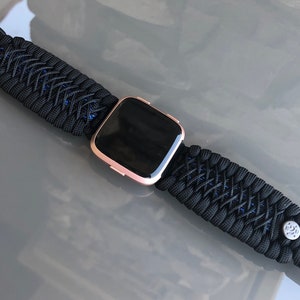 Puede incluir: Una correa de reloj de paracord negra con un smartwatch de oro rosa. La correa tiene un dise&ntilde;o tejido con detalles azules y una hebilla negra.