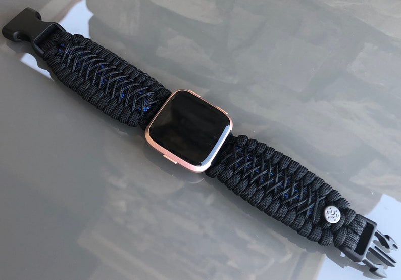 Puede incluir: Una correa de reloj de paracord negra con un smartwatch de oro rosa. La correa tiene un dise&ntilde;o tejido con detalles azules y una hebilla negra.