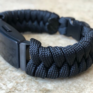 Op de afbeelding: Zwart paracord armband met een donkergrijze metalen sluiting en een geweven ontwerp. De armband heeft een rechthoekig metalen stuk en een veilige gespsluiting. Dit accessoire is geschikt voor buitenactiviteiten.