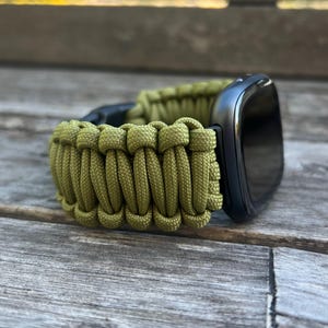 Pode incluir: Uma pulseira de relógio de paracord verde com um mostrador preto.