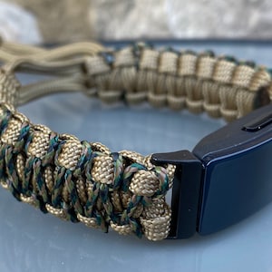 May include: A tan and green paracord bracelet with a black fitness tracker attached. The bracelet is made with a woven pattern.