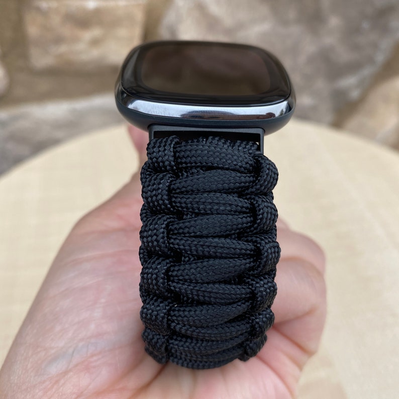 May include: Black paracord watch band with a black smartwatch face. The band is made of woven paracord.