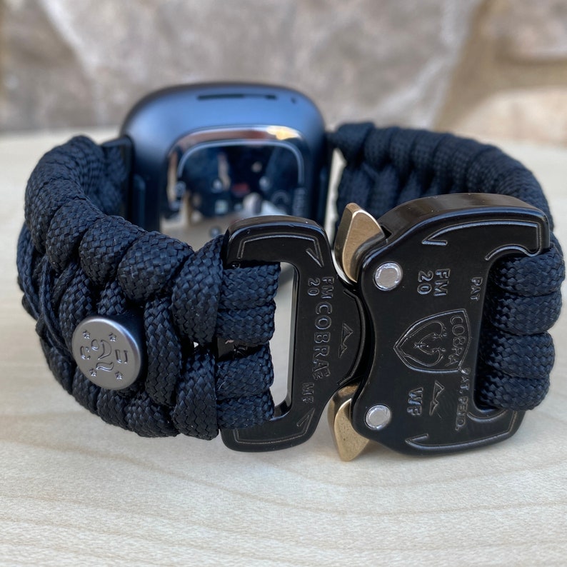 May include: Black paracord watch band with a black metal buckle and a gold metal clasp. The buckle has the text "FM COBRA MF 20" and "FM 20" printed on it. The clasp has the text "COBRA PAT PEND" printed on it.