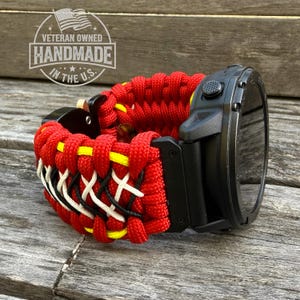 Puede incluir: Un reloj inteligente negro con una correa de paracord roja, amarilla, negra y blanca. La correa está hecha a mano y presenta un diseño tejido. El reloj se muestra sobre una superficie de madera. El texto en la imagen dice "Veteran Owned Handmade in the U.S."