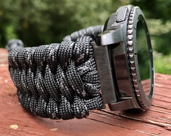 May include: A black smartwatch with a black paracord strap. The watch face is black with white markings. The watch has a rotating bezel.