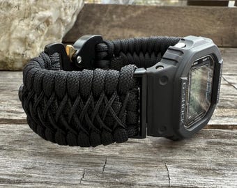 Puede incluir: Un reloj G-Shock negro con una correa de paracord negra. La correa está tejida con un patrón de diamante y tiene una hebilla negra.
