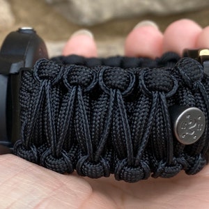 May include: A black paracord watch band featuring a complex woven pattern with a series of interconnected knots. A small, circular, metallic silver-toned button with an engraved logo and star accents is attached to the side of the woven cord structure.