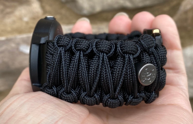May include: A black paracord watch band featuring a complex woven pattern with a series of interconnected knots. A small, circular, metallic silver-toned button with an engraved logo and star accents is attached to the side of the woven cord structure.