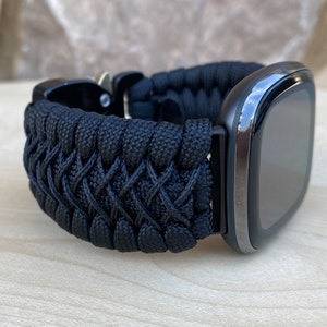 May include: A black paracord watch strap with a black buckle. The strap is woven in a criss-cross pattern.