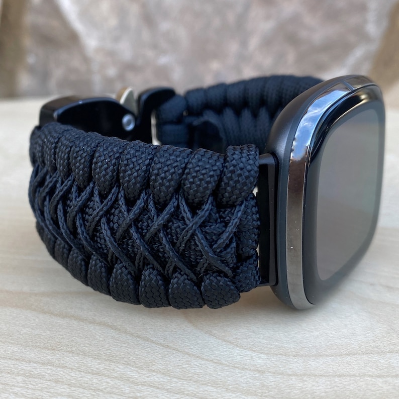 May include: A black paracord watch strap with a black buckle. The strap is woven in a criss-cross pattern.