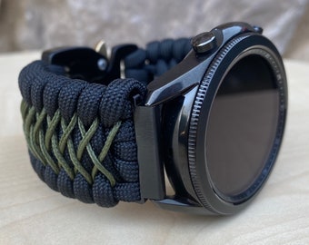 May include: Black smartwatch with a black and green paracord strap. The watch face is black and the strap is woven with a criss-cross pattern.
