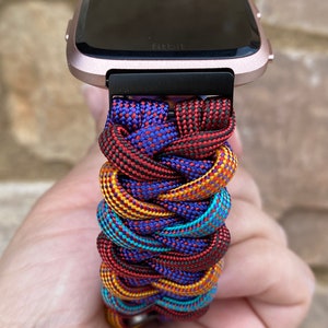 Puede incluir: Un reloj inteligente Fitbit de color rosa dorado con una banda de paracord trenzada de colores. La banda est&aacute; hecha de paracord rojo, amarillo, azul y negro.
