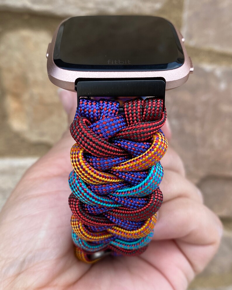 Puede incluir: Un reloj inteligente Fitbit de color rosa dorado con una banda de paracord trenzada de colores. La banda est&aacute; hecha de paracord rojo, amarillo, azul y negro.