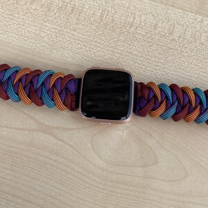Puede incluir: Una pulsera de reloj trenzada en morado, marr&oacute;n y azul con una hebilla negra y un reloj inteligente de oro rosa.