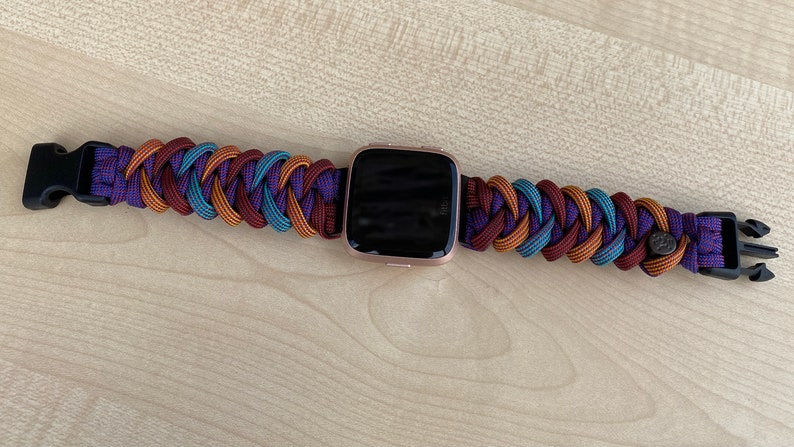 Puede incluir: Una pulsera de reloj trenzada en morado, marr&oacute;n y azul con una hebilla negra y un reloj inteligente de oro rosa.
