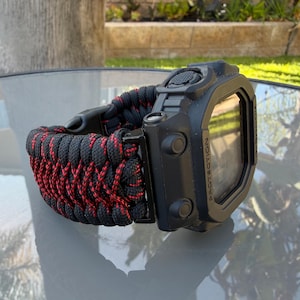 May include: A black digital watch with a rectangular case featuring the word PROTECTION on the side. The watch has a thick, woven paracord strap in a black and red pattern with a black plastic buckle closure and side control buttons.