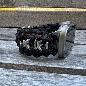May include: A black and red paracord watch band with silver metal accents. The band is woven with a pattern of nuts and bolts. The band is attached to a silver Apple Watch.