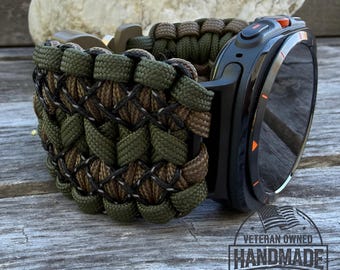 May include: A black smartwatch with a camouflage paracord band. The band features a woven pattern in shades of green, brown, and black. The watch has an orange button and a digital display. The image includes a "Veteran Owned Handmade in the U.S." logo.