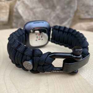 May include: Black paracord watch band with a black metal clasp. The band is made of woven paracord and has a metal buckle with the word "CORDING" engraved on it.