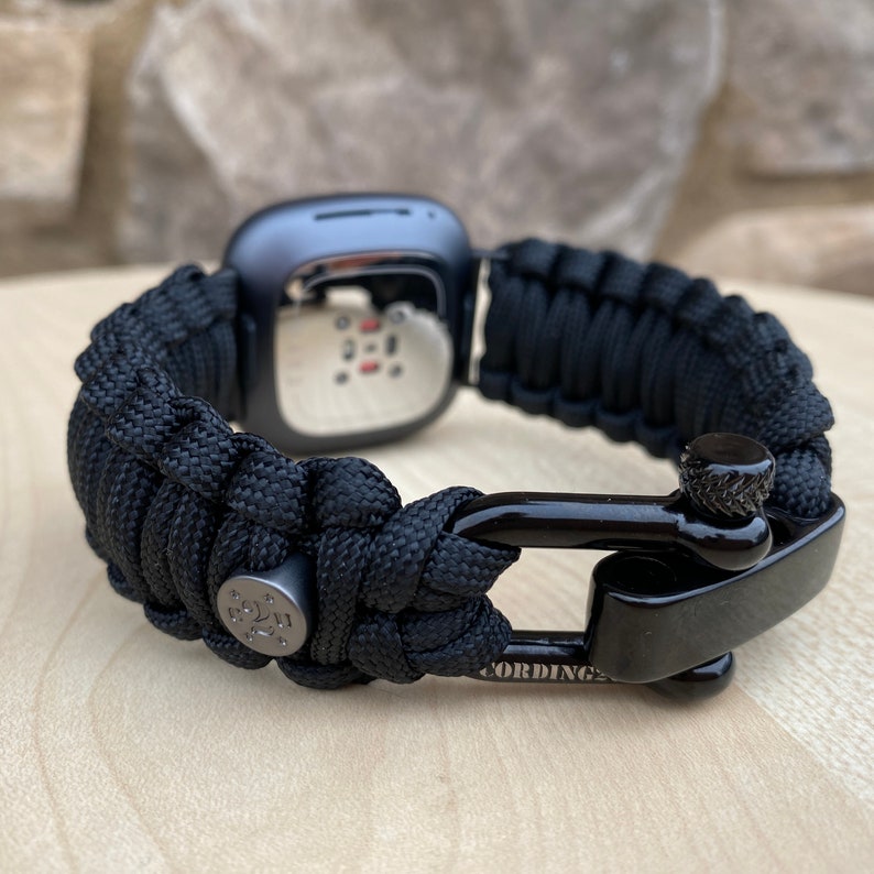 May include: Black paracord watch band with a black metal clasp. The band is made of woven paracord and has a metal buckle with the word "CORDING" engraved on it.