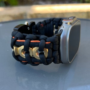 May include: A black and orange woven watch band with gold accents. The band is made of paracord and has a metal buckle. The band is designed to fit a smartwatch.