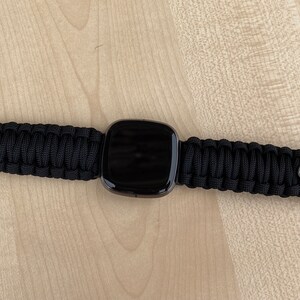 May include: A black smartwatch with a black paracord strap. The strap has a metal clasp on each end.