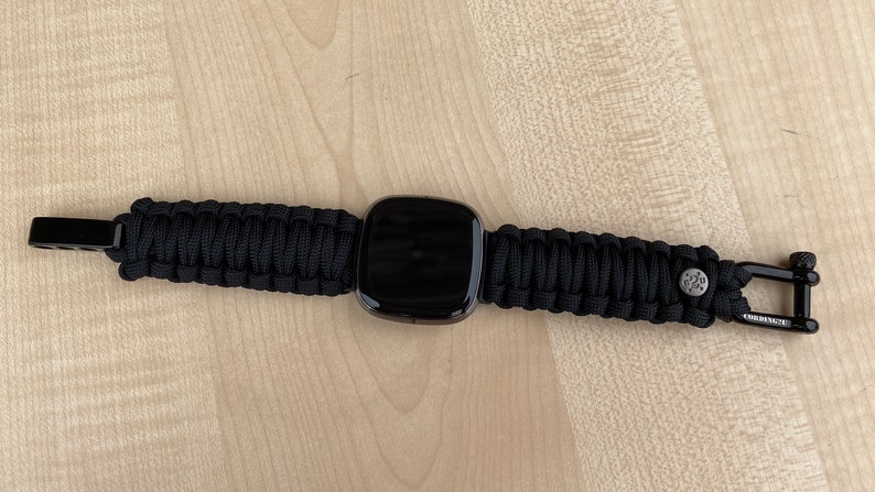 May include: A black smartwatch with a black paracord strap. The strap has a metal clasp on each end.